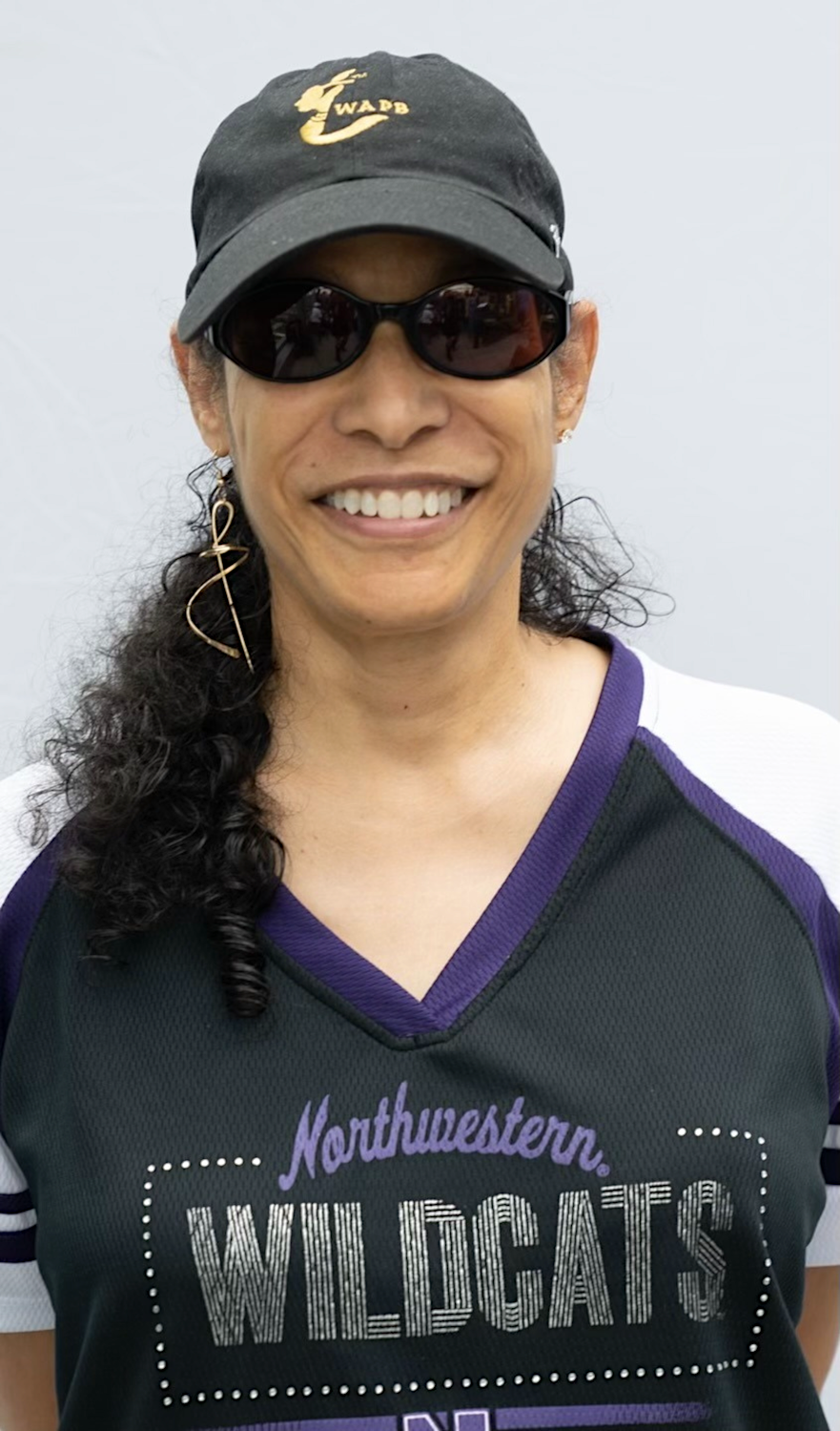 An image of Crista Noel, Founder of WAPB and mentor to Youth International's team. She has long, curly black hair and wears a black WAPB cap, dark sunglasses and a Northwestern University Wildcats jersey.