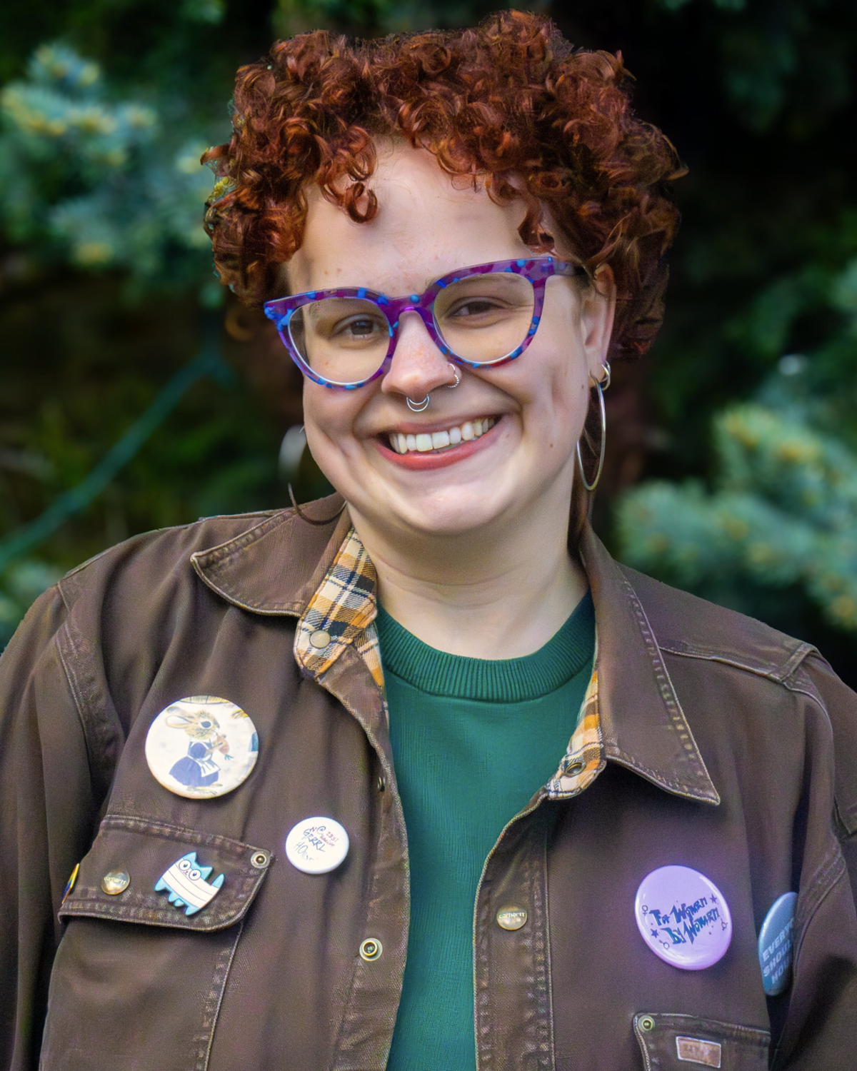 An image of Edie Danielle, Co-founder of Youth International. They have short, curly red hair and wear a green shirt under a brown jacket, adorned with various pin buttons.
